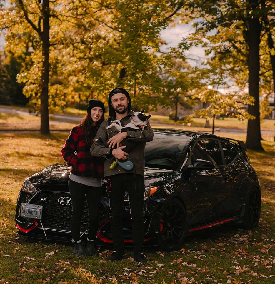 A car parked on grass with fallen leaves, while a man holding a dog and a woman standing beside him pose in front of it.